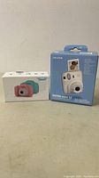 Boxes of the two cameras, showing Fujifilm Instax Mini 12 and pink children's camera packaging