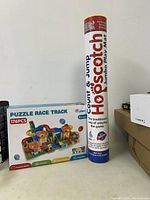 Photo shows Hopscotch Count & Jump Jumbo Play Mat packed in a tall cylindrical tube next to Puzzle Race Track box with colorful race track design on front.