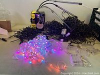 Multiple bundles of black cord LED string lights and one bundle of white cord multicolor LED string lights illuminated with pink, blue, purple and white colors gathered on table with two boxes.