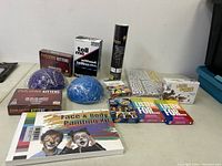 View of all items on table: Exploding Kittens (2 boxes), Tell Me game, glow sticks tube, Little Nutters card game, blue and purple round wrapped items, stack of stickers, two Listen For games, UNO cards, face and body painting kit.