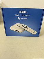 Front of packaging showing Mecool KM2 Android TV box and remote control image with brand and 'Hey Google' voice assistant mention.