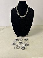 Chain necklace displayed on black bust and eight packaged men's rings laid on table.