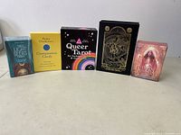 Front view of all five card decks lined up: "The Light Seers Tarot," "Compassion Cards," "Queer Tarot," "Gold Foil Tarot," and "The Divine Feminine Oracle"