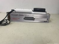 Hair straightener comb placed on its box, showing product and packaging approximately from the front and side angle.