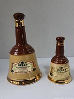 Photo showing two Bell's Blended Scotch Whisky ceramic bell-shaped bottles, one larger and one smaller, with brown tops and gold accents, on a neutral background.
