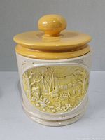 Front view of ceramic cookie jar with yellow lid and embossed rural scene