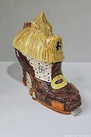 Side view of ceramic boot-shaped cookie jar with thatched roof design resembling a cottage, showing textured brown boot and beige-yellow roof with chimney.