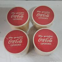 Four rolls of disposable paper coasters with red tops labeled 'The genuine Coca-Cola Experience.'
