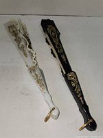 Two closed hand fans, one white and one black, both featuring ornate gold and floral decorative patterns on the sticks and guards.