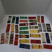 Top-down view of about 30 vintage matchbooks laid flat in rows showing a variety of colors and printed advertisements.
