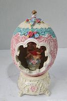 Front view of the music ornament showing pastel egg-shaped enclosure with floral designs and cherub inside