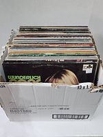Box filled with many vinyl records, some covers visible but mostly packed tightly.