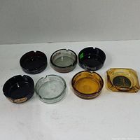Seven vintage ashtrays arranged in two rows on a white background including black, clear, amber, and smoked amber glass ashtrays with various branding and shapes.