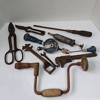 Photo showing vintage hand tools including a hand drill, scissors, wrenches, rasp, and screwdriver on white surface.