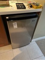 Bar fridge positioned under countertop showing front closed door with brand label and sticker.