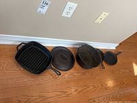 Four cast iron cookware pieces arranged on hardwood floor in natural light including three skillets of various shapes and one pot with lid.