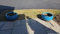 Two blue glazed ceramic planters positioned outside on paving stones and grass, showing top and side views with soil inside each.