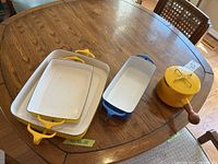 Photo showing all four items: sauce pot with lid and wood handle, yellow loaf pan, yellow and blue baking pans on a wooden table.