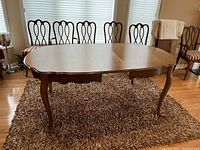 Wooden oval dining table with five wooden chairs in a living room setting on a shaggy rug. Table has one 12-inch leaf inserted to extend surface area.