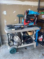 Front and side view of beige and green plastic rolling garden workbench holding various garden tools, potting mix bags, and cast resin decorative logs stored underneath.