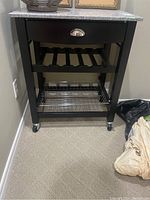 Full front view of the black wood kitchen cart showing the stone top, drawer with silver handle, wine rack slats, wire basket, and caster wheels.