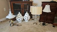 Full view of two brass metal bedside table lamps with milk glass hurricane shades, two white ceramic urn candleholders with pierced clover pattern tops, and a metal desk lamp with beige rectangular shade visible from left to right on carpet.