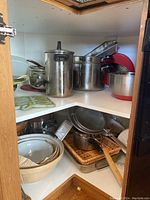 Corner cabinet shelf showing multiple stainless steel pots, a tall pot with lid, and some kitchen utensils and pans stacked around