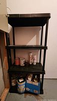 Photo of black plastic shelving with 4 shelves, holding assorted paint cans and spray paint on a concrete floor and wall corner.