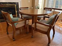 Wide angle showing round wooden pedestal table with 4 matching chairs, wicker cane backs, upholstered seats, on castors, positioned on hardwood floor near fireplace and windows.