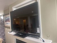 Full view of black Sony flat panel television mounted on a white shelf with TV stand and attached power cord, showing reflective screen.