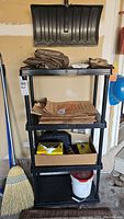 Front view of the 4-level black plastic utility shelf with various miscellaneous items on and inside the shelves, including folded tarps, cardboard boxes, and a white bucket underneath.