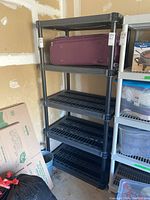 Full view of black plastic utility shelf with a maroon suitcase on top shelf, showing five shelves in total, measuring approximately 36 x 24 x 74 inches.