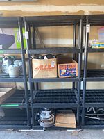 Front view of black plastic modular utility shelf showing four shelves with grid pattern and tubular posts. Two cardboard boxes and other garage items seen around it.