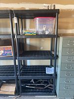 Full view of black plastic modular utility shelf showing five open slatted shelves. Shelf is empty except for a clear plastic bin on top shelf and some other storage boxes around it, contents not included.