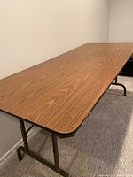 Photo showing the 6 foot folding table with wood grain laminate finish on the top and black metal folding legs supporting it.