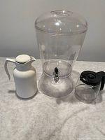 Photo showing three assorted drink holders: a white insulated pitcher, a large clear beverage dispenser with spigot, and a small glass coffee carafe with black handle and lid.