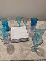 Photo shows a collection of assorted plastic kitchenware including stacks of white square plastic plates and multiple translucent plastic drinking glasses and wine glasses in various colors.