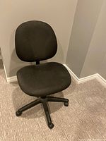 Photo of a black office chair with a cushioned seat and backrest, five wheels on the base, placed on a carpeted floor in a corner of a room.