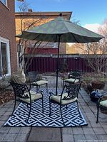 Four black wrought iron chairs with pale green cushions arranged around a green leaf-pattern patio umbrella on a black and white geometric rug, showing umbrella and stand.