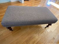 Photo showing the rectangular cloth ottoman with fabric upholstery, dark wooden turned legs, and caster wheels placed on a hardwood floor.