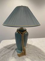 Photo of green ceramic table lamp with pleated green shade and gold tassel wrapped around the lamp base.