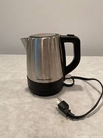 Photo of a small Hamilton Beach electric kettle made of stainless steel with a black plastic handle, base, and power cord.