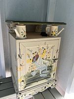 Front view of a small wooden cupboard hand painted with a floral scene including a white chair with a red bird, yellow flowers and greenery, two hand painted small watering cans on top.