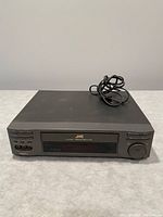 Front view of JVC VHS player showing the front-loading slot, control buttons including play, stop, rewind, fast forward, and a large dial on the right, power cord coiled on top.