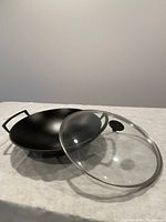 Black cast iron wok with two side handles and accompanying clear glass lid laid on a light surface