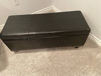Photo of a black leather rectangular storage ottoman with top open for storage, showing nailhead trim and caster wheels.