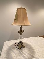 Light beige table lamp with decorative tassel on shade, ornate metal and glass base shown on white surface against neutral wall.