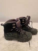 Photo shows a pair of black Merrell men's hiking boots size 10 with lace-up fronts and rugged soles placed on a surface against a plain wall.