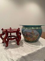 Photo shows a decorative porcelain fishbowl with colorful painted scenes of birds and fish beside a carved red wooden base stand on a white cloth surface.
