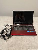Front view of red Samsung laptop open showing keyboard and screen, with power adapter and cord placed beside it on a flat surface.